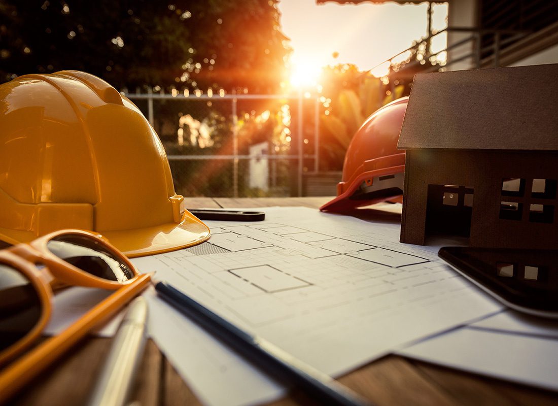 Insurance by Industry - Closeup View of New Home Blueprints a Model Home and Contractor Hard Hats on a Wooden Desk Outside at Sunset