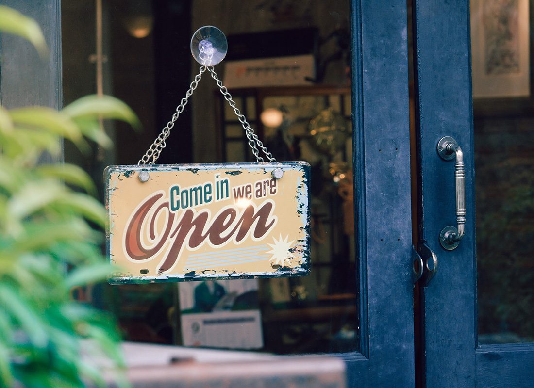 Small Business Online Quoting - Close Up of an Come In We Are Open Sign Hanging on a Window Next to the Glass Door of a Small Business on a Nice Day